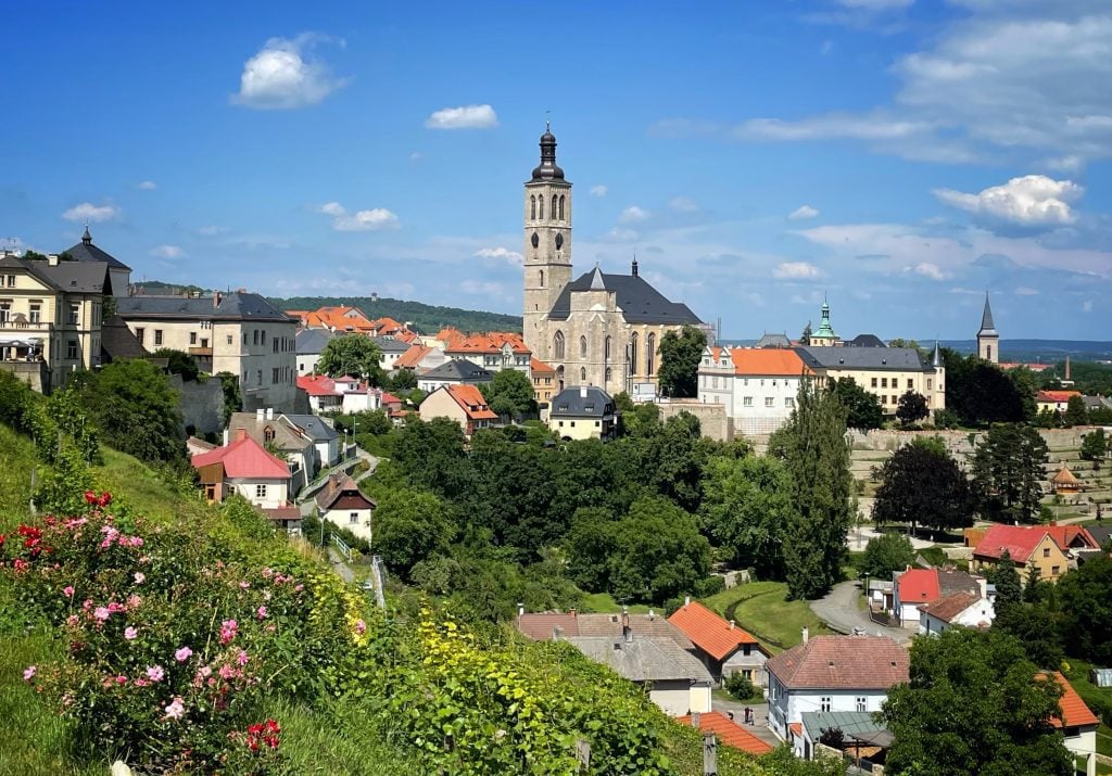 The Bohemian town of Kutna Hora underneath a blue sky with clouds. You see a church tower, lots of small buildings with orange roofs, and a hillside topped with pink and red flowers.