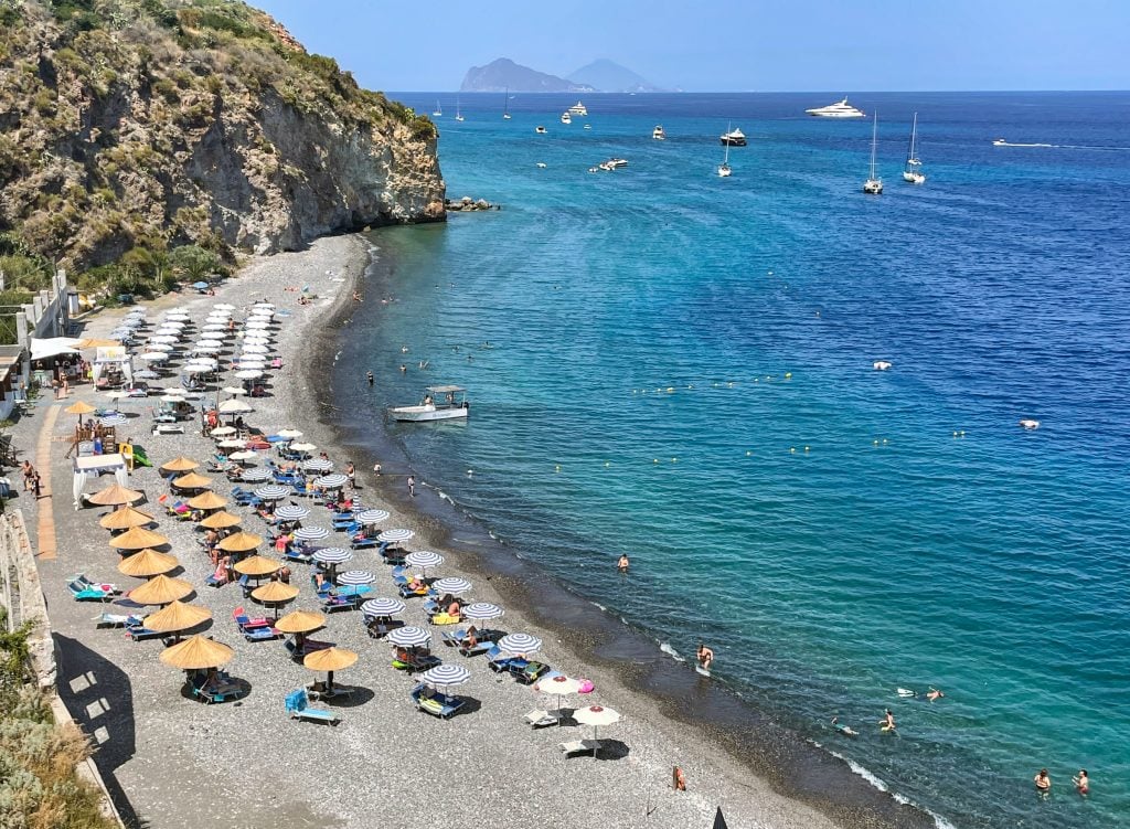 A view from above of the gray rocky beach of Lipari, rows of umbrellas, and a bright blue sea.