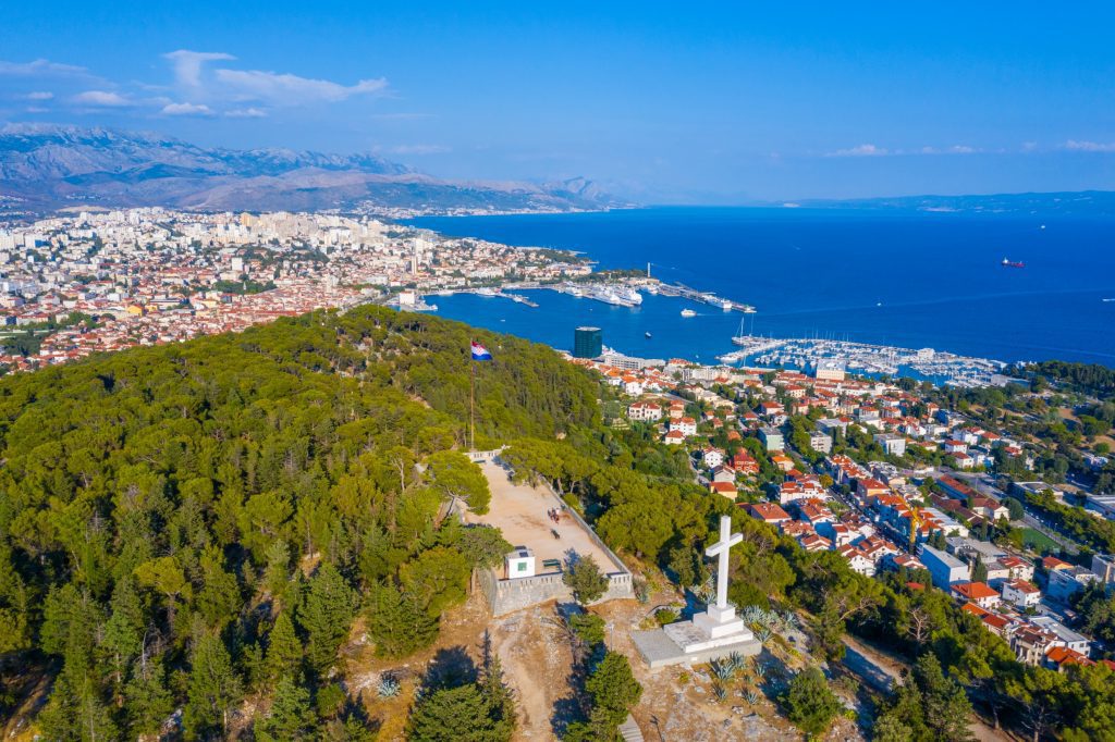 An aerial view of a big forested hill with a giant stone cross and a Croatian flag on it, the city of Split way down in the background.
