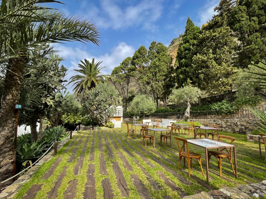 The grounds of Ngonia Bay: an open green area with tons of cactus and tree landscaping. The ground had boards interspersed with green grass, and there are several sets of tables topped with Sicilian tiles and wooden chairs.