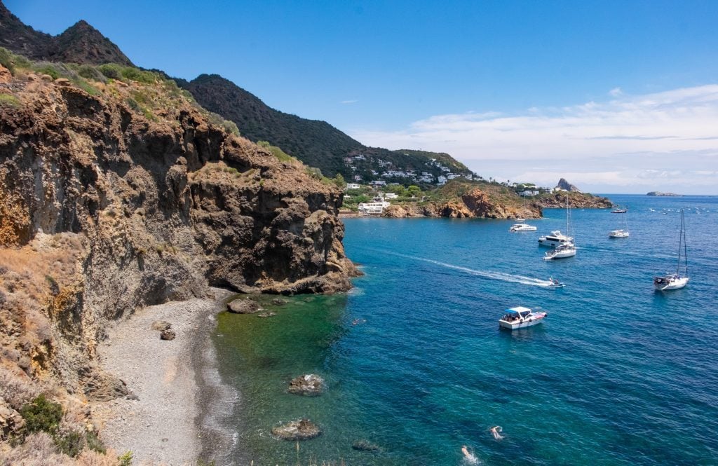 View of tall brown cliffs of Stromboli, small gray pebbly beaches, and several sailboats docked in the bright blue-green water.