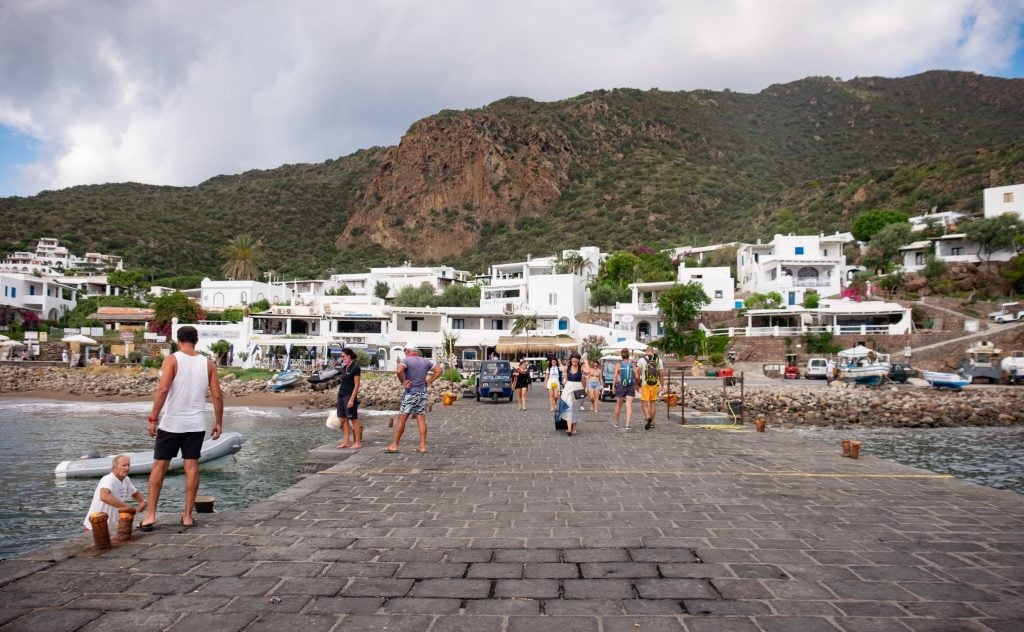 The dock of Panarea: a long gray brick pier leading to a small town filled with blocky white buildings. People dragging suitcases to the dock to get on the ferry.