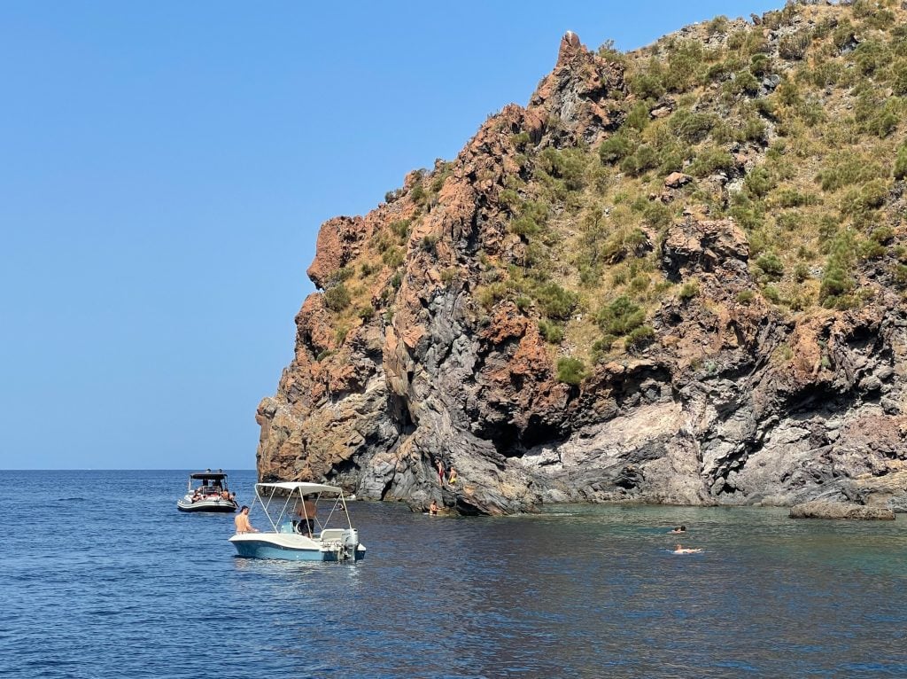 On the red-brown coast of Vulcano, you see a small pool that is clearer and greener than the other ocean water, and some people swimming and boating near it.