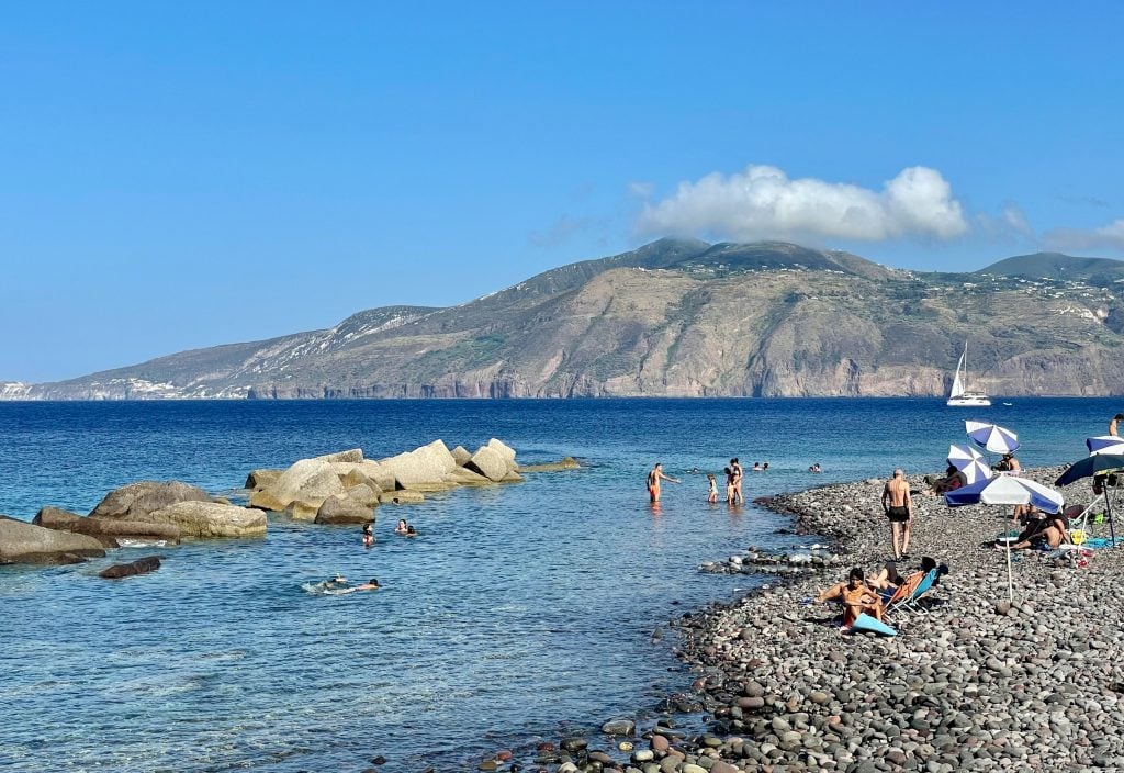 Punta Lingua, a small beach with large but not huge gray rocks leading into clear shallow water, a few kids and adults in the water. Lipari island is in the distance.