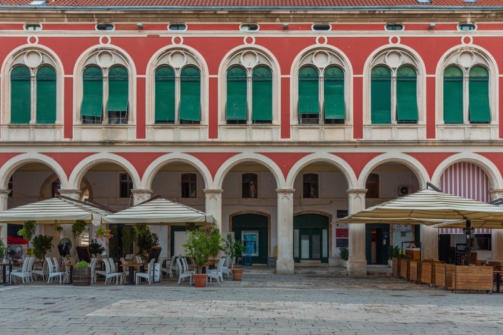 Republic square with its light red walls, emerald green shutters, and rows of porticoes along the bottom, cafe tables in front.