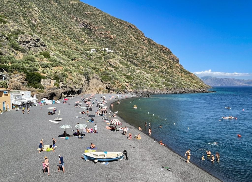 Rinnella Beach with a few dozen people on it. You see small gray stones, very calm teal water with almost no waves. Lots of kids there.