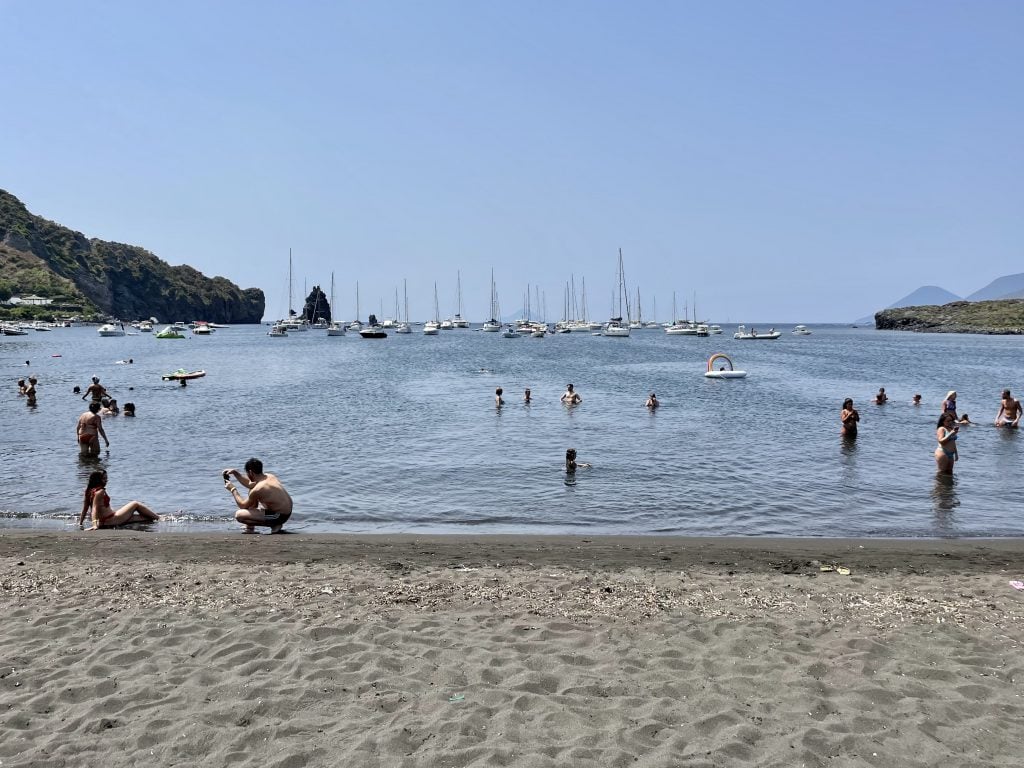 Sabbie Nere: a boring-looking gray sand beach with several dozen people in the water, several of them taking photos of themselves or each other.
