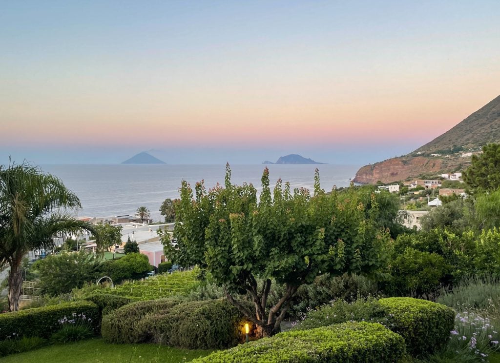 Views of green landscaping of Salina, cliffs in the distance, and you see the islands of Stromboli and Panarea peeking above the coastline as the sun sets.