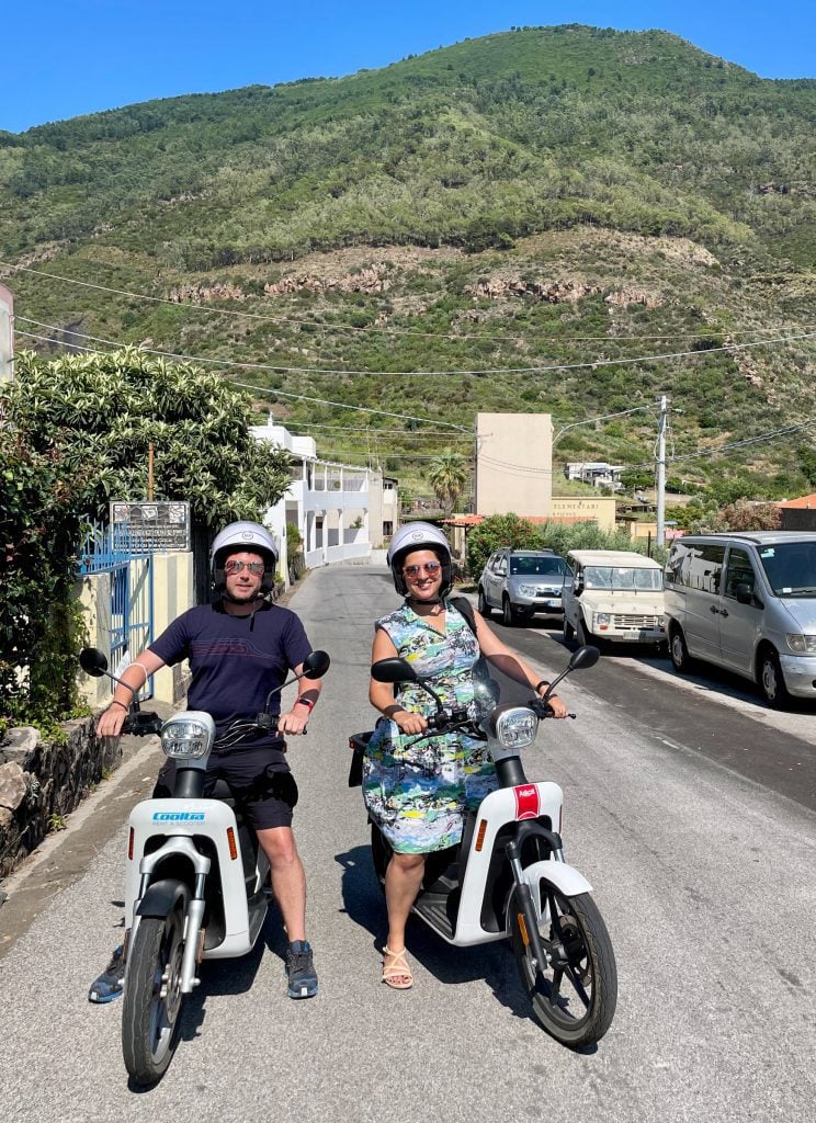 Kate and Charlie side by side on white electric motorbikes, a green mountain rising behind them. Kate wears a 50s-style green and white patterned housewife dress.