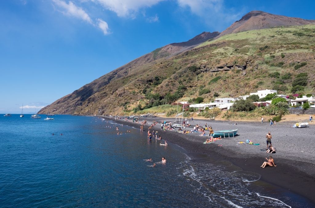 The island of Stromboli, with dozens of people sunning themselves on a black sand beach and playing in the blue water, the volcano rising behind them.