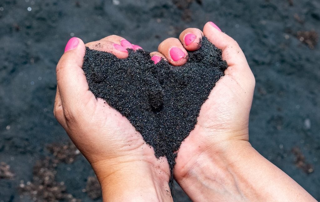 Kate's hands (with bright pink fingernails) holding a handful of sparkling black Stromboli sand, her hands forming the shape of a heart.