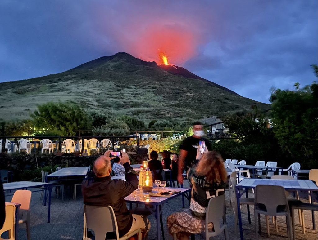 The volcano of Stromboli, spewing an explosion of bright red lava in a darkening sky. In the foreground, there are outdoor tables at Osservatorio restaurant and people have their phones out photographing the lava.