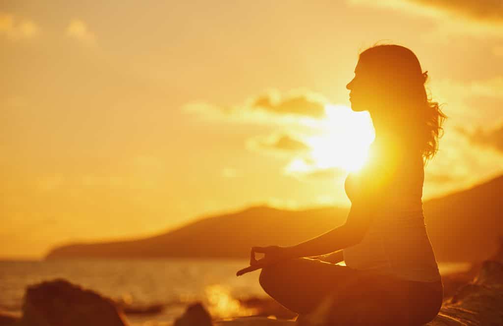 A woman doing yoga in lotus pose by the water in front of an orange sunset.