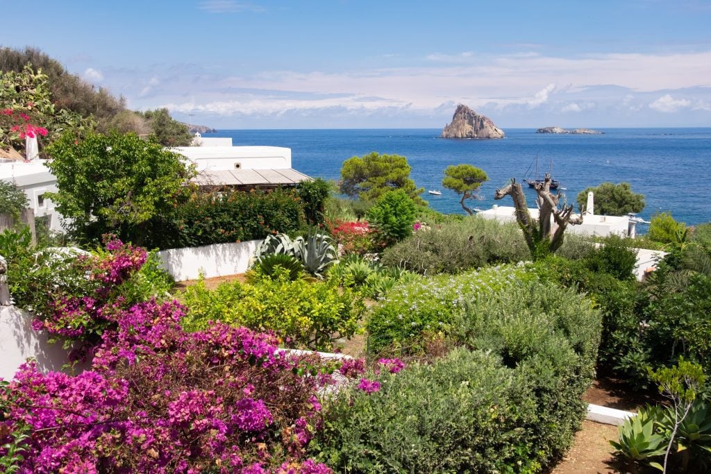 Views of a garden bursting with green and purple trees and bushes, overlooking the ocean, a triangular rock formation bursting out from the ocean.