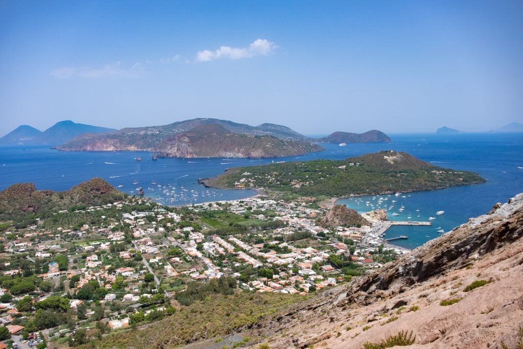 An insane view of Vulcano island and several islands in the distance. Vulcano is bright green and filled with white homes. You see lots of boats in the water.