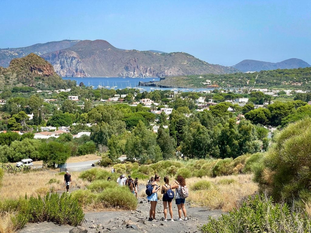 The view from the beginning of climbing Vulcano, with three teenage girls wearing backpacks and shorts having a conversation. In the background you see the green landscape, studded with white blocky homes, leading to the blue ocean, topped with white sailboats, and more islands in the distance.