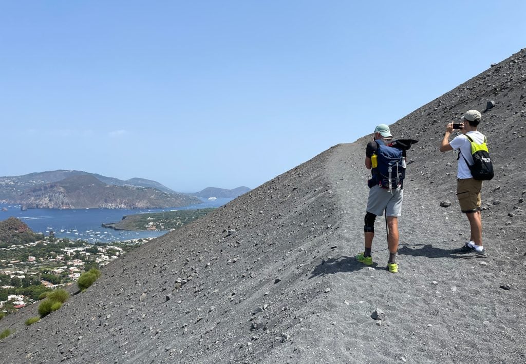 Two hikers taking photos of islands in the distance while climbing a sandy gray path on Vulcano.