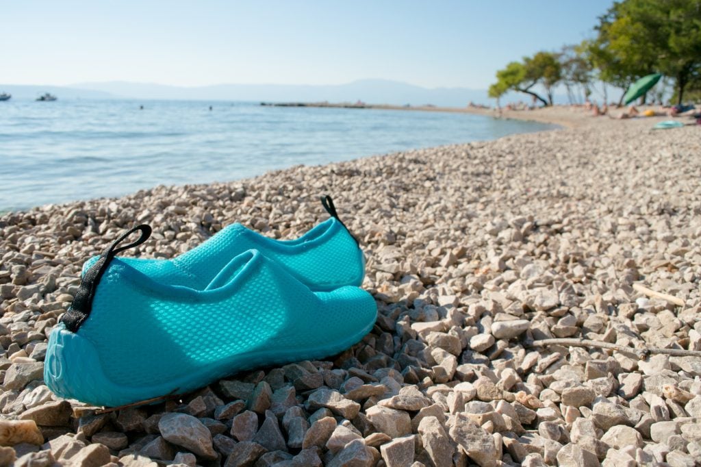 A pair of turquoise water shoes sitting on a gray stoney beach.