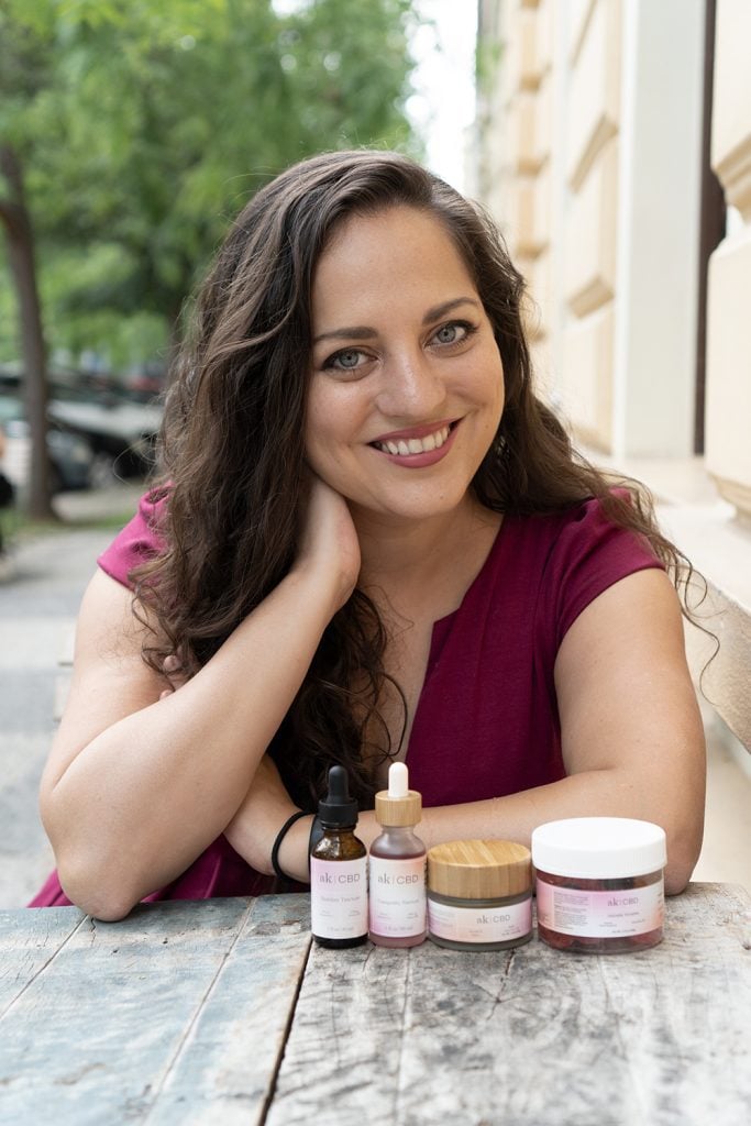 Kate sits at an outdoor cafe table and smiles at the camera, two bottles of AK CBD tinctures, one jar of soothing salve, and one jar of gummies in front of her.