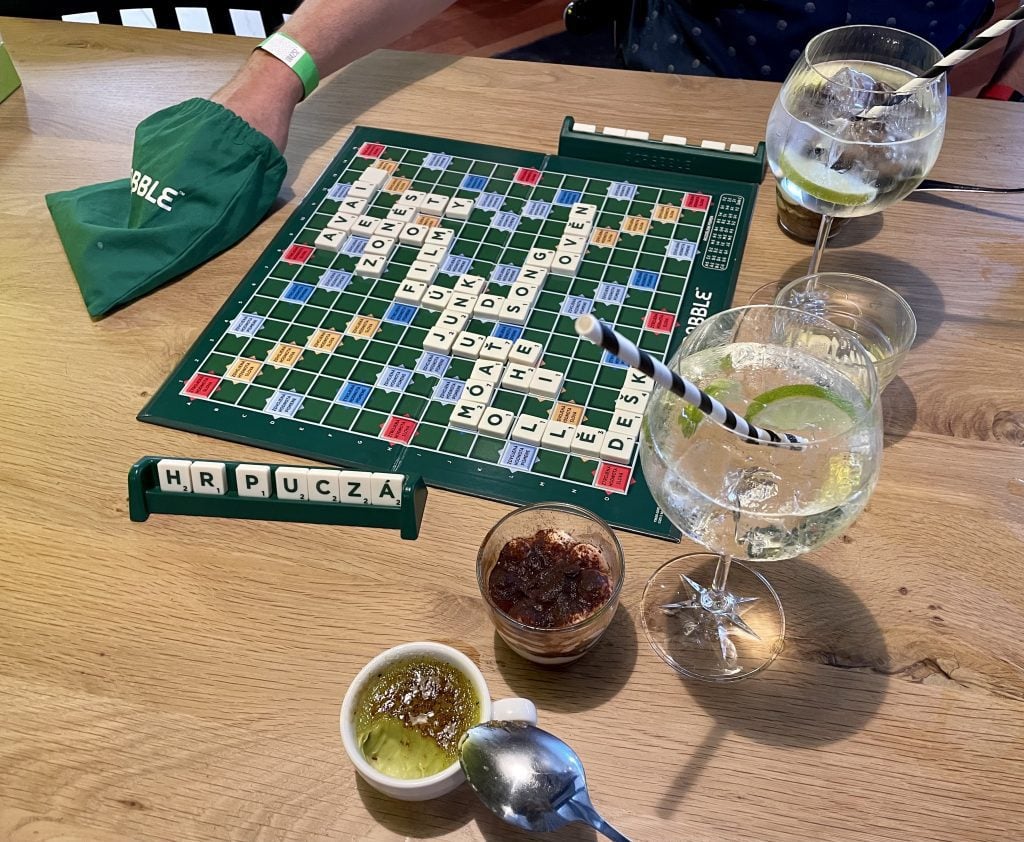 A Scrabble board that is clearly Czech with Czech letters, plus two cocktails, a pistachio creme brûlée, and tiramisu.