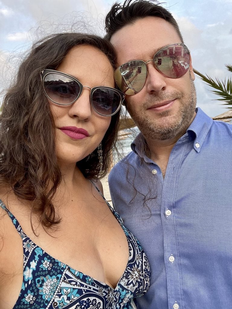 Kate and Charlie take a smoldering selfie, both wearing sunglasses and smiling gently. Kate's sunglasses, a gift from Charlie, are black and cat's eye shaped, outlined in gold, with the trademark gold T-shape of Tom Ford on the outer corners. Kate wears a low cut spaghetti strapped blue and white paisley patterned dress. Charlie wears reflective aviator sunglasses (surprise: they're actually Kate's) and a blue button-down shirt.