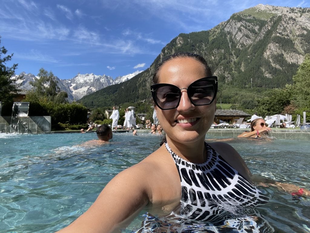 Kate takes a selfie and smiles in a black and white patterned bathing suit and sunglasses. She's in a pool and in the background you see green mountains, then taller snow-covered gray mountains.