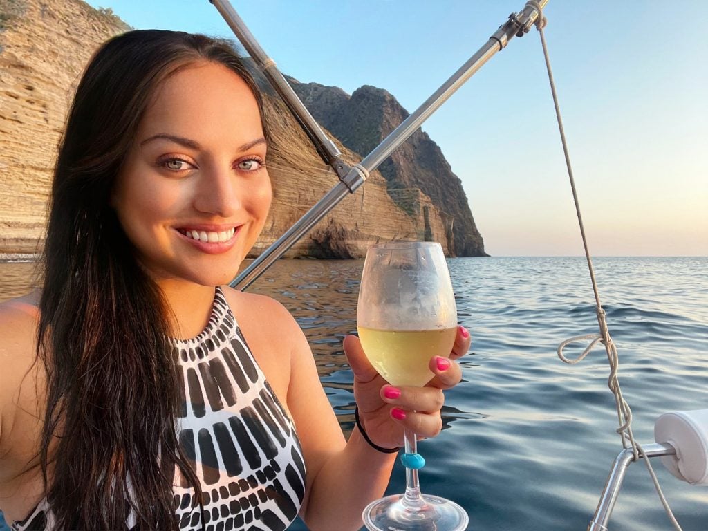 Kate smiles and wears a black and white patterned bathing suit while sitting on a boat. She's holding a glass of white wine and behind her you see tall striped cliffs and blue water.