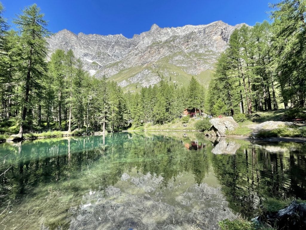 A lake with a mirror-like reflection of gray mountains and green pine trees, underneath a blue sky. There's a wooden cabin on shore.