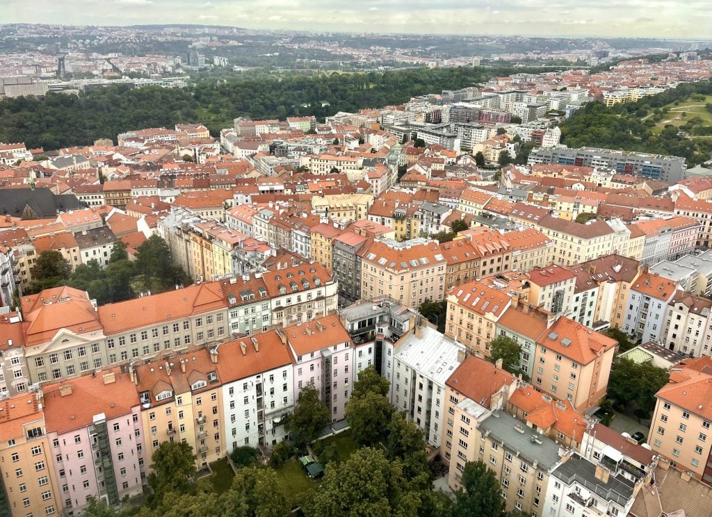 A view of Prague from the top of the TV tower: you see grids of pink, yellow, orange, and white-colored apartment buildings surrounding courtyards, all with orange roofs.