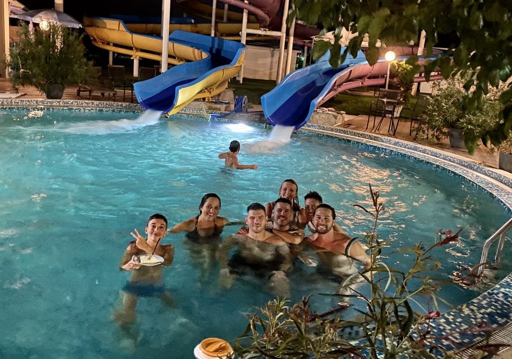 Kate and five of her friends, plus two teenage boys, in a pool into which two waterslides are feeding. One of the boys is eating a slice of cake in the pool!