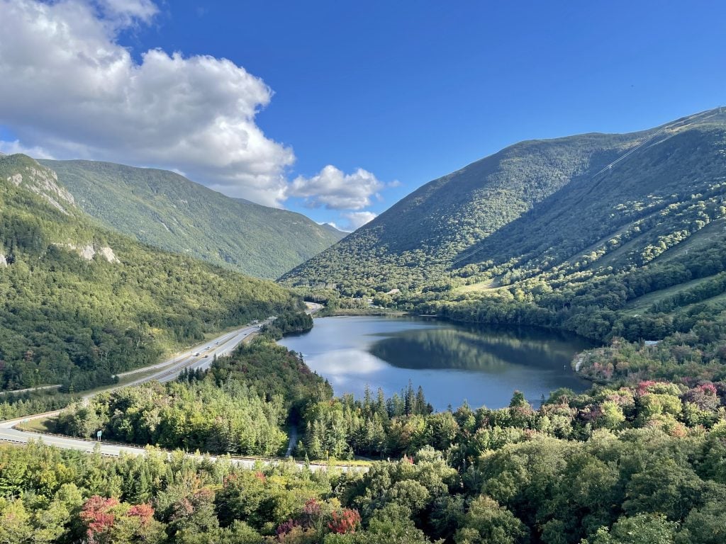 View through the middle of a valley surrounded by green mountains, a small highway snaking by the left side and a calm navy blue glassy lake in the middle. A few of the trees are starting to turn red.