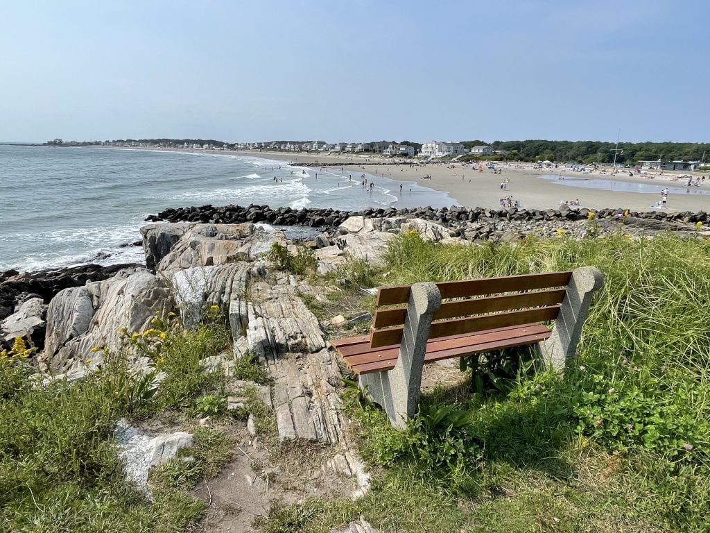 A weathered wooden and concrete bench placed among tall grasses on a rocky cliff overlooking a long beach in Rye, New Hampshire.