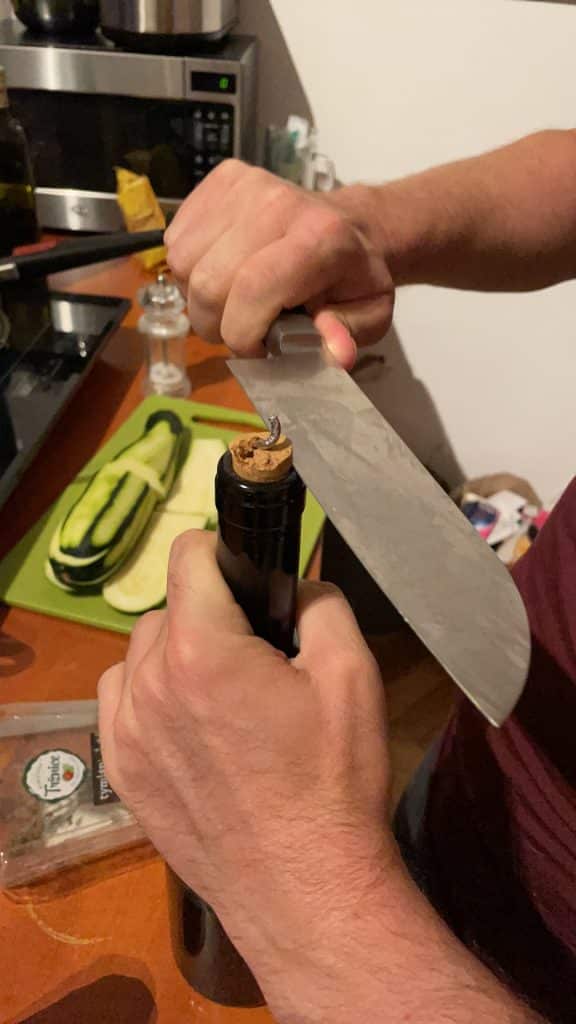 A wine bottle with the cork partway out, the metal spiral wine opener lodged in it. Charlie's hands are trying to cut the cork out with a giant butcher knife.