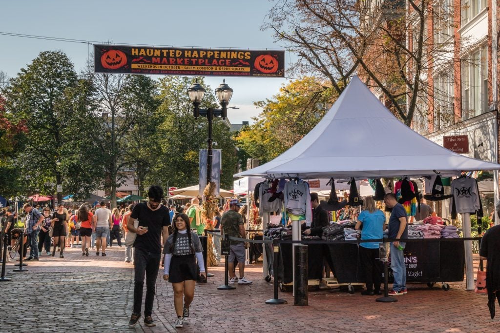 A couple walking on the cobblestone street in front of a street fair with stalls selling Salem sweatshirts and witch hats.