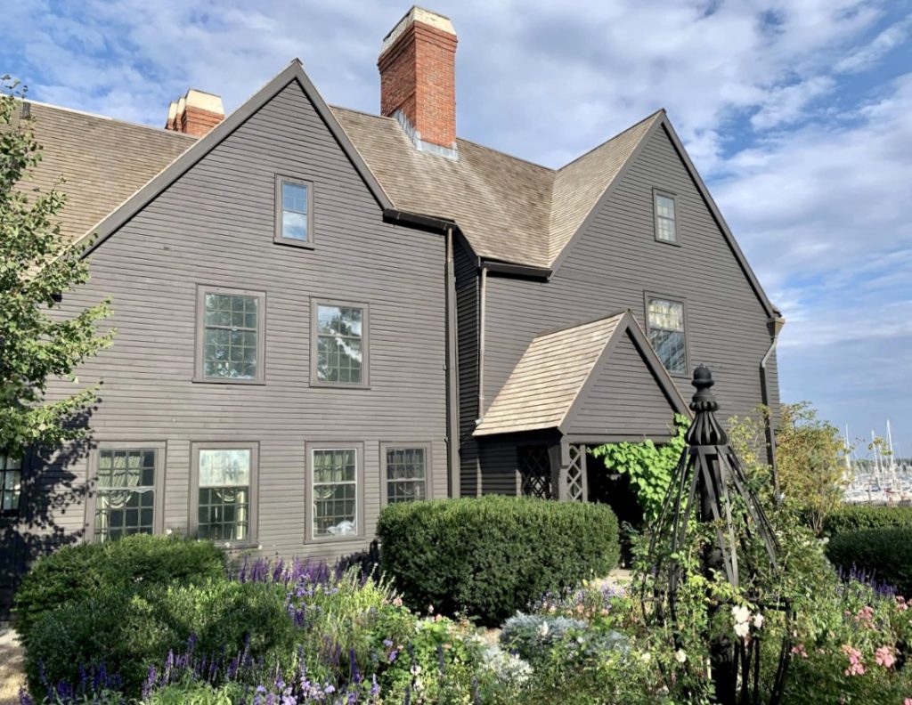 The dark gray House of the Seven Gables with several points on its rooftop. In front of it is a garden of purple and white flowers.