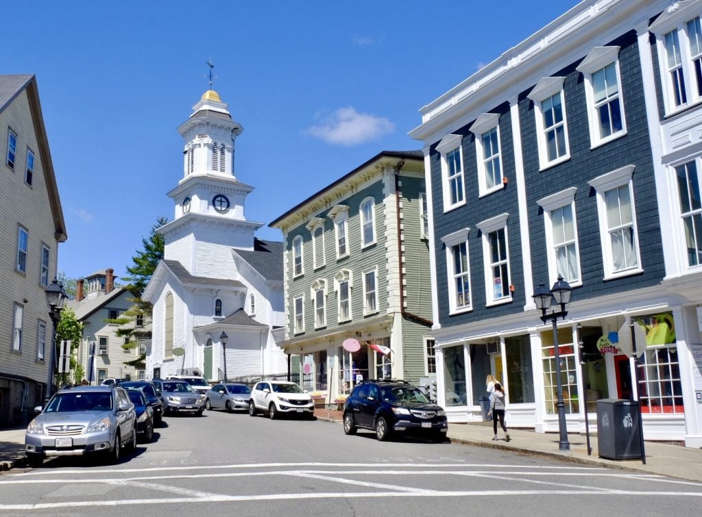A small street in Marblehead with a tall white church next to old-fashioned but well-manicured buildings painted pea green and dark blue.