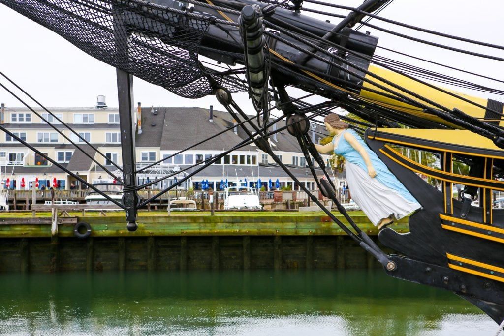 The front of a ship with a wooden woman in a dress leading the way, set in a green harbor.