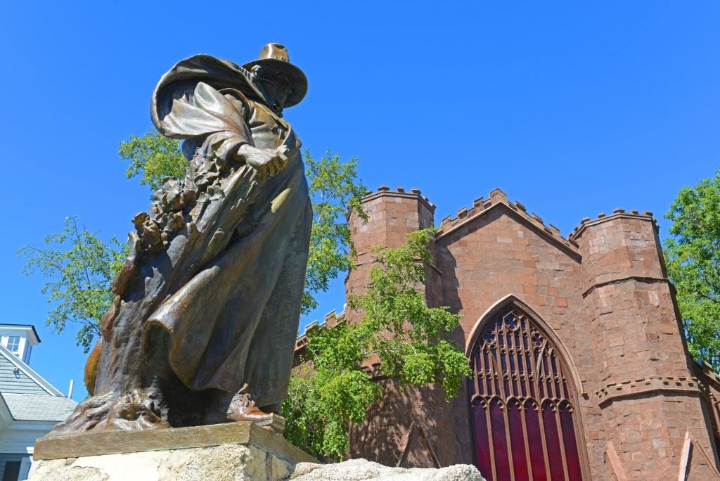 A close-up of a statue of a man in 1600s garb in a robe and wide-brimmed hat. In the background, the reddish clay-colored Salem Witch Museum, with a big stained glass window in the front.