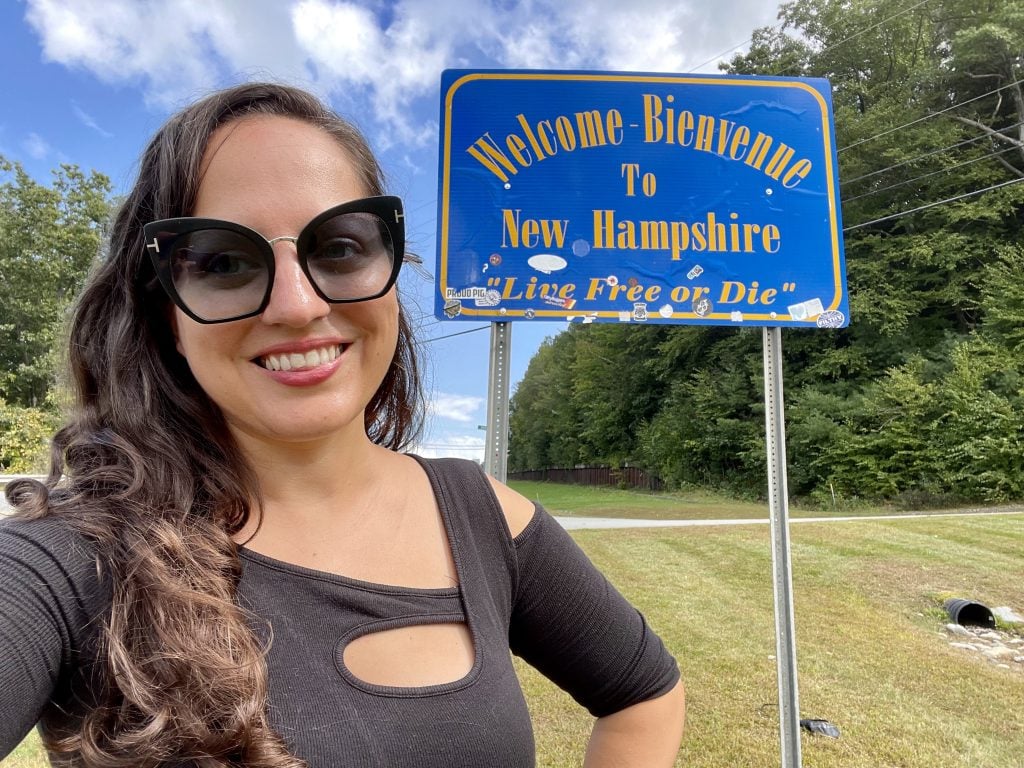 Kate wears sunglasses and a black shirt with cutouts and stands in front of a side reading "Welcome-Bienvenue to New Hampshire. Live Free or Die."