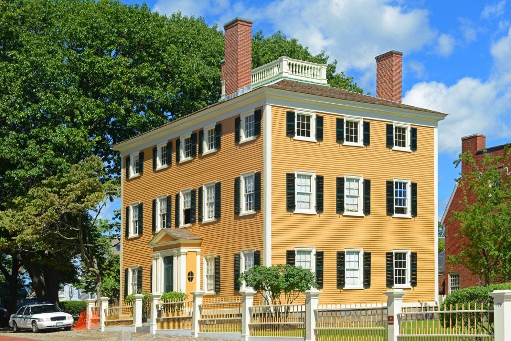 A large, square, mustard-yellow house with three floors and lots of square windows with black shutters.