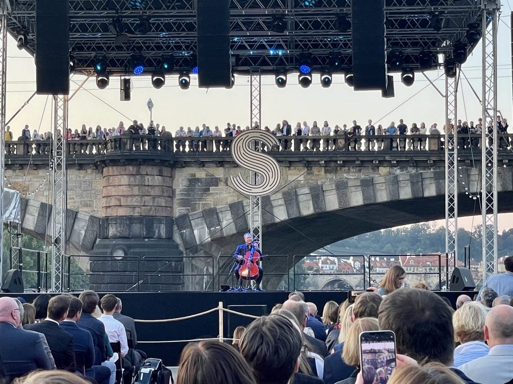 Yo-Yo Ma sitting in the center of an outdoor stage with his cello. Behind him is a bridge lined with people watching him perform.