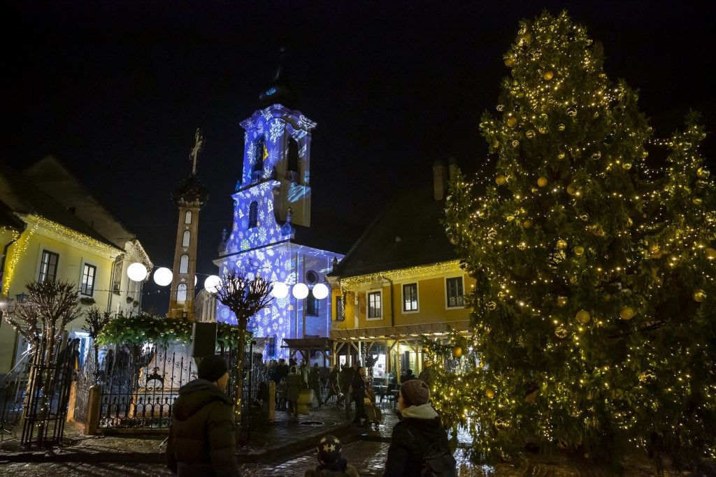 A shot of the Christmas Market at night, with a Christmas tree covered in gold bulbs and the small white church having a white-on-blue snowflake pattern projected onto it.
