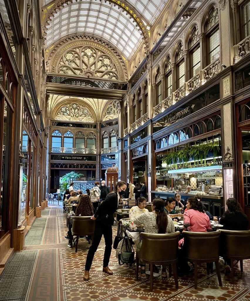 An cafe in Budapest with high glassed-in ceilings, ornate woodwork, and wildly patterned tiles in warm colors.