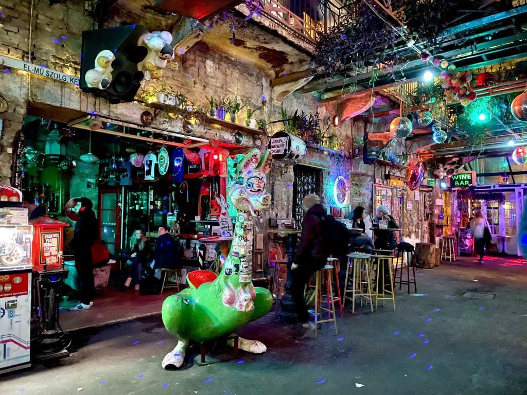 The wild Szimpla ruin bar: it's dark, neon lights are everywhere, several disco balls and plants hang from the ceiling, a dinosaur cartoon statue is covered with bumper stickers, and pretty much everything is thrown on the walls, as people sit on bar stools and enjoy drinks.