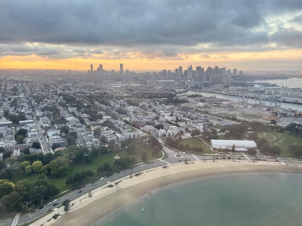 A cloudy sunset over the city skyline of Boston. You see a beach and gray water along the edge.