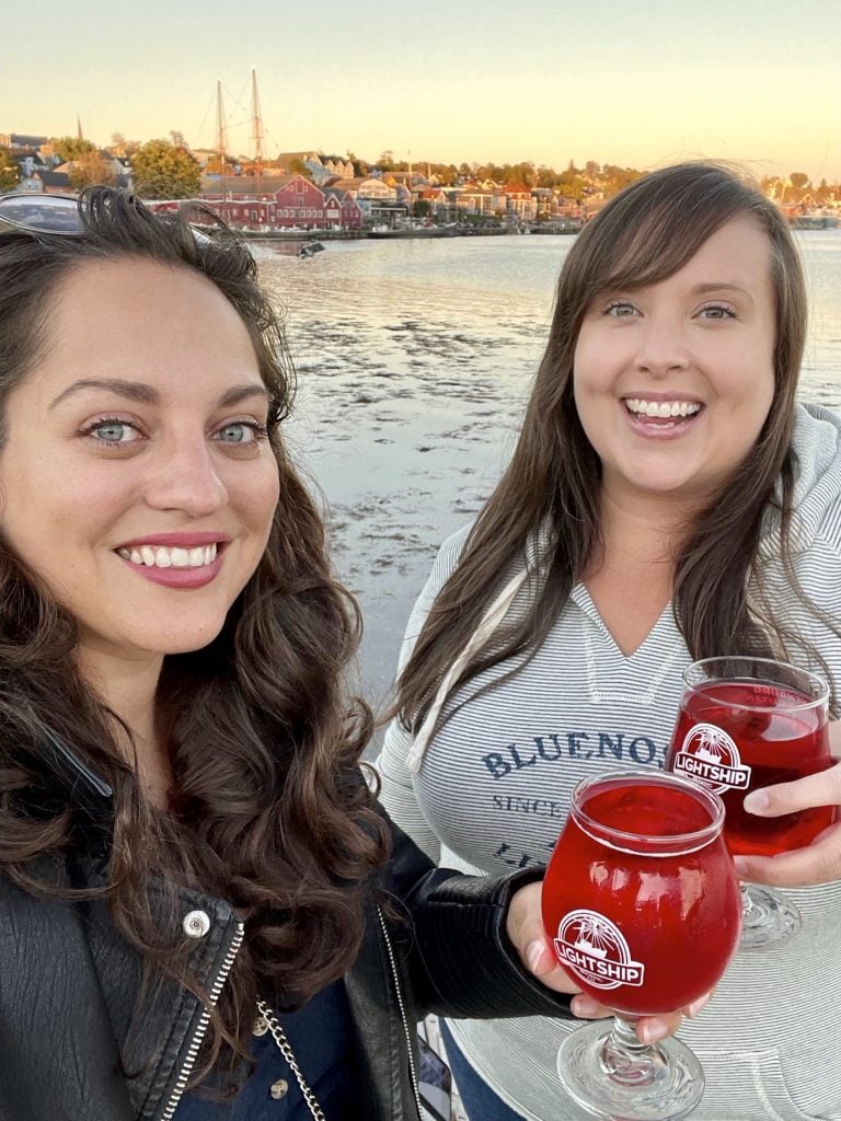 Kate and Cailin posing for a selfie with glasses of bright red cranberry orange cider. Lunenberg harbor is in the background.