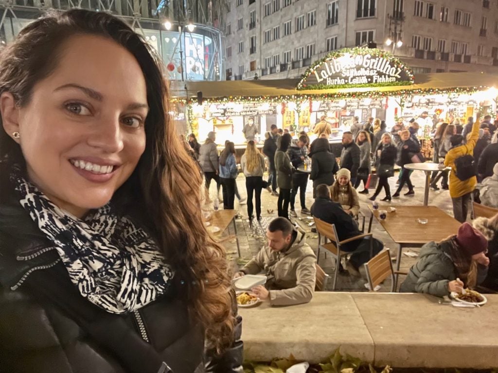 A selfie of Kate smiling in front of a market stall covered with pine garlands and ornaments; people eating at tables in front of it.