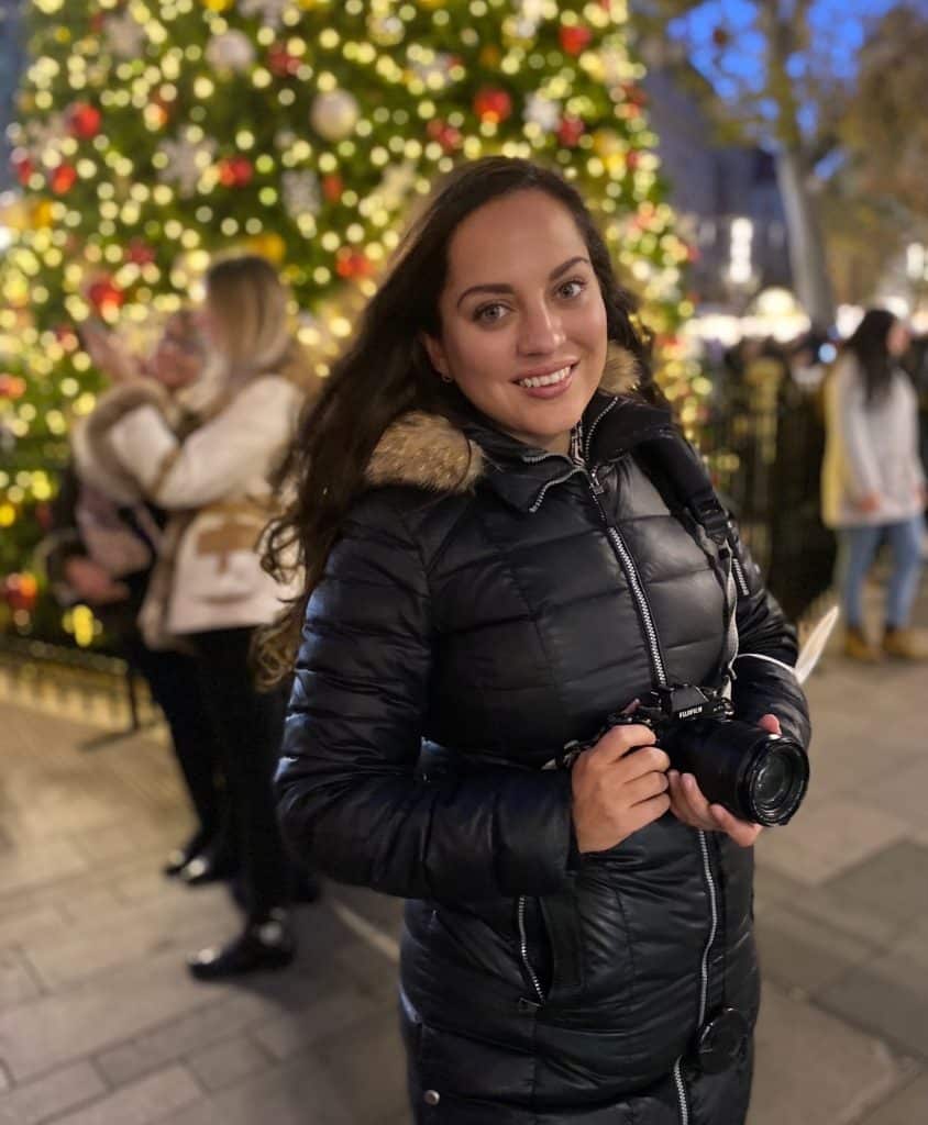 Kate smiling, wearing a long black coat and carrying a black Fuji camera, posing in front of a Christmas tree.