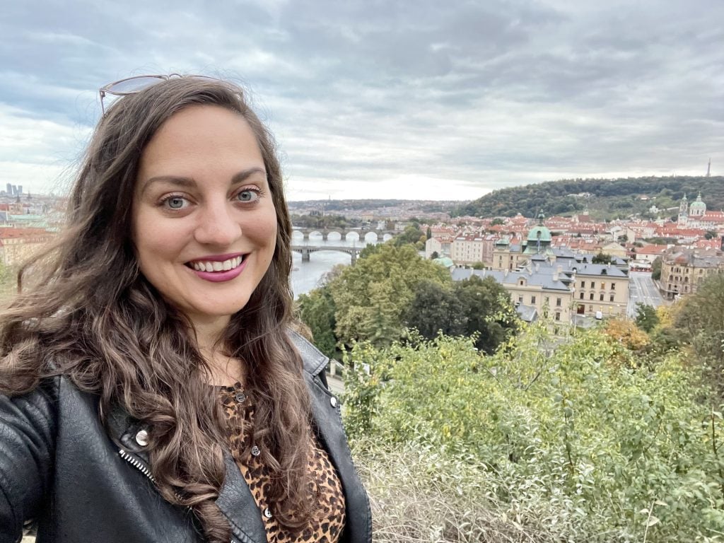 Kate takes a selfie on a cloudy Prague day in front of the river in Prague, several bridges on it. She wears a black leather jacket and a leopard-print blouse.