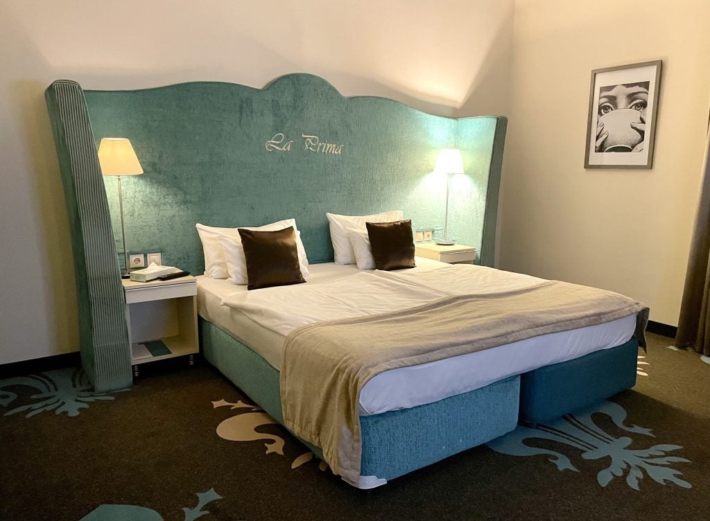 A bed with an enormous wraparound aqua-colored headboard reading La Prima, a tall white lamp on each white side table.